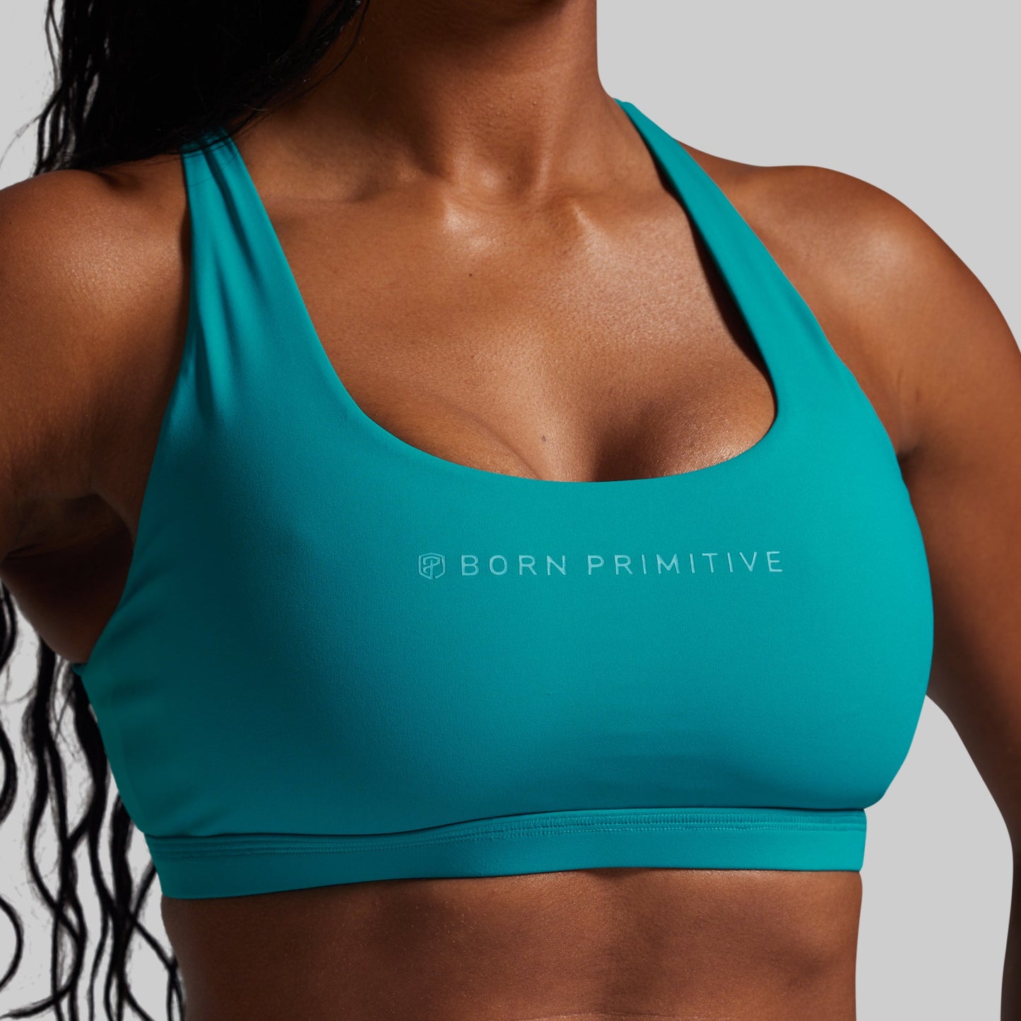 Teal sports bra with 'Born Primitive' logo worn by a person on a gray background