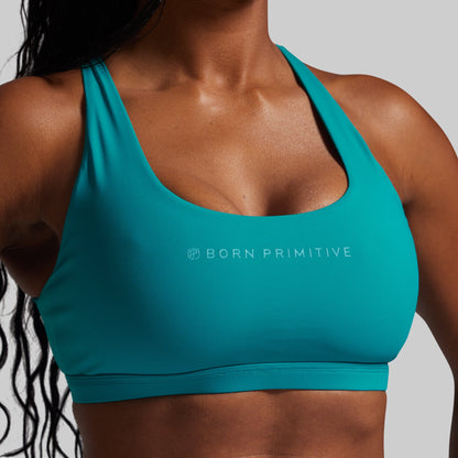 Teal sports bra with 'Born Primitive' logo worn by a person on a gray background