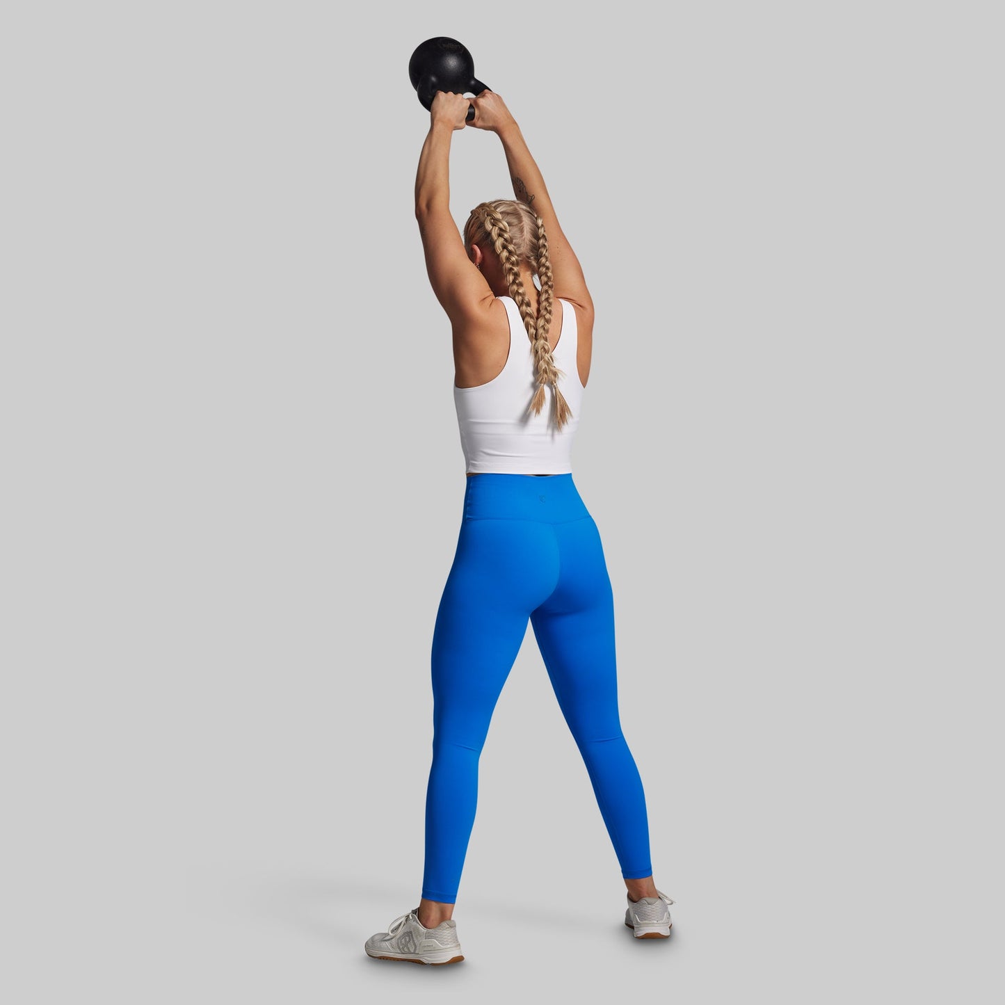 Person lifting a kettlebell wearing blue leggings and a white tank top on a gray background