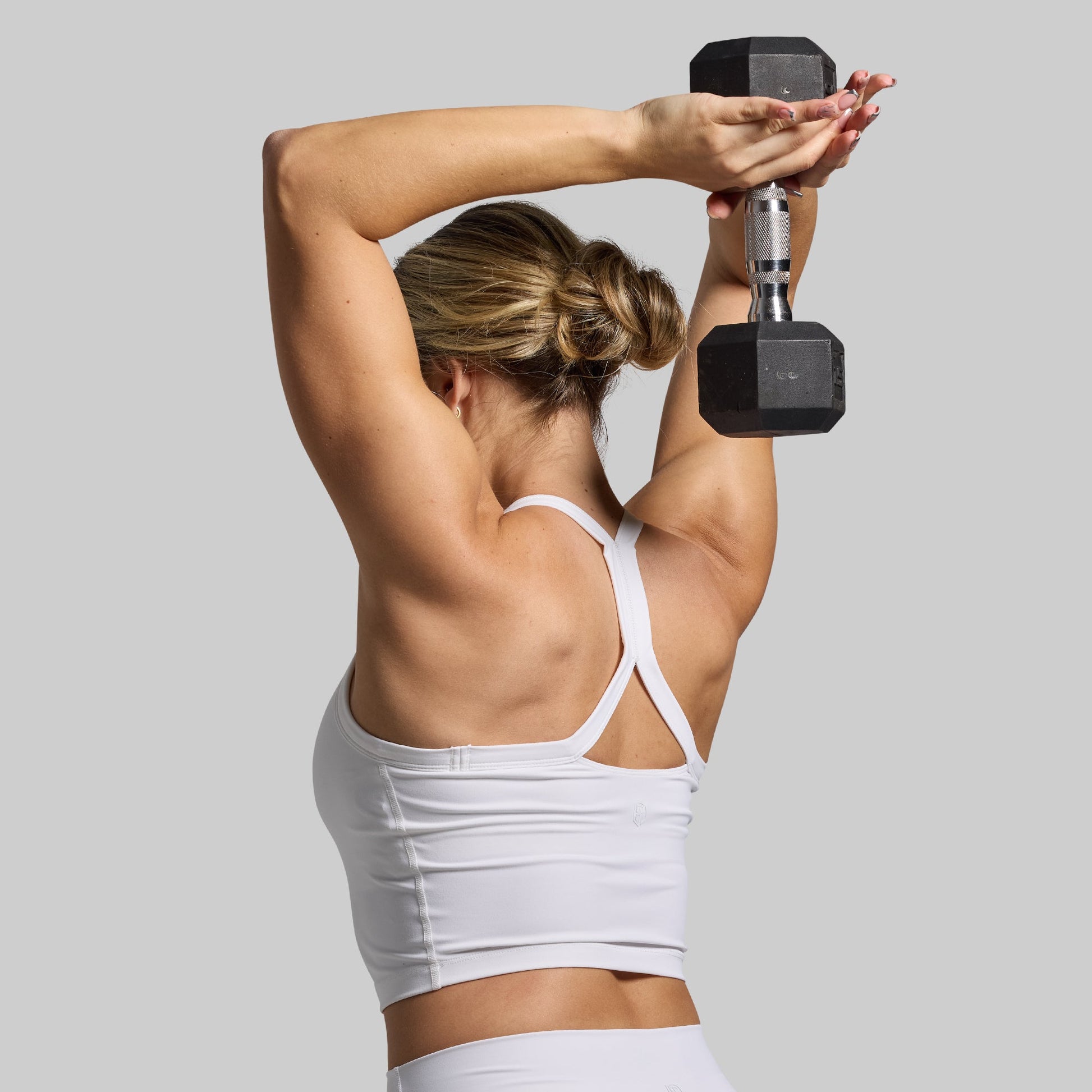 A person is seen from the back wearing a white sports bra, with their arms raised holding a dumbbell above their head.