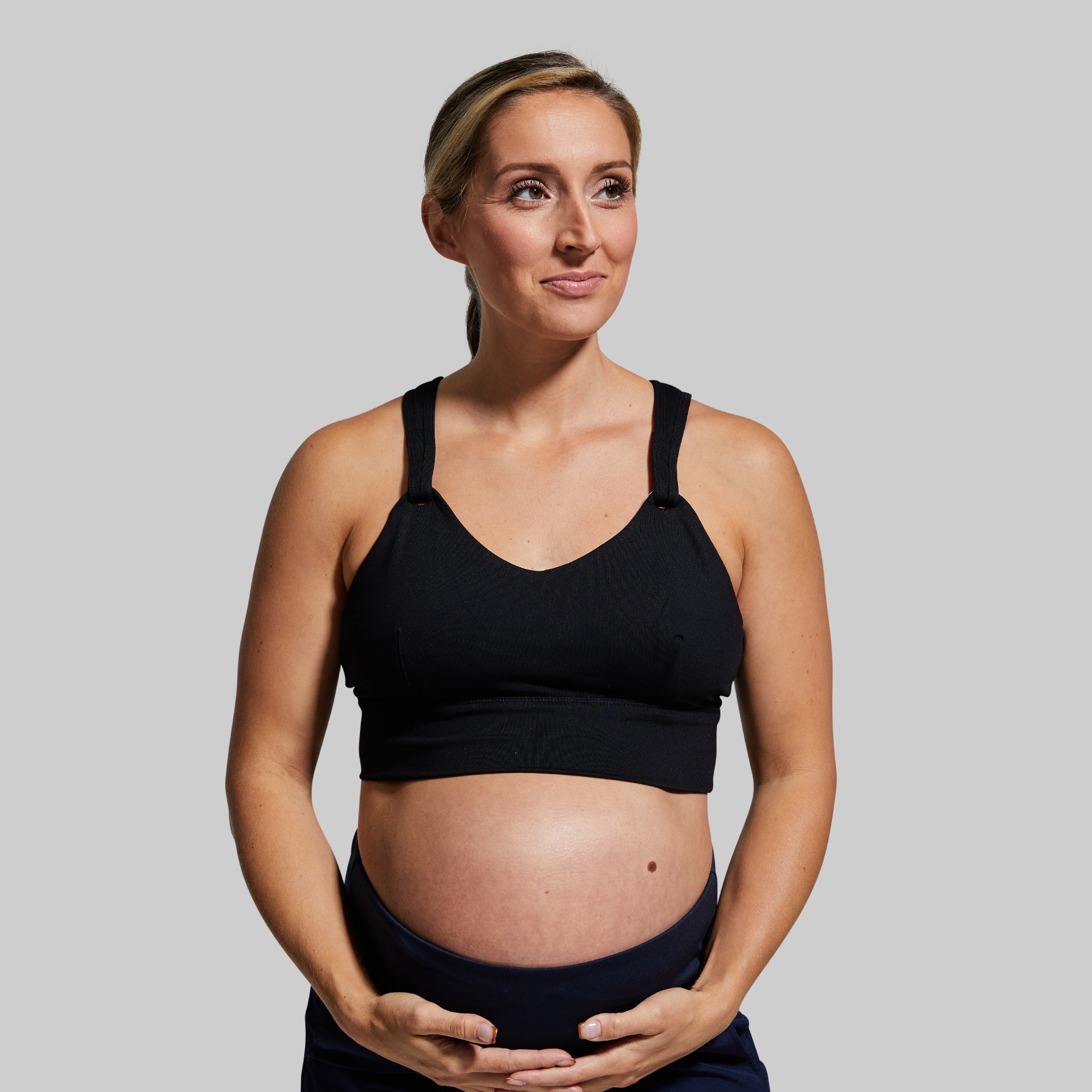 Pregnant woman wearing a black sports bra and black leggings against a plain background