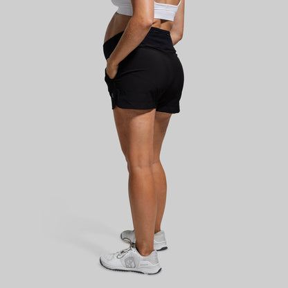 Pregnant person wearing black maternity athletic shorts and white sneakers on a plain background