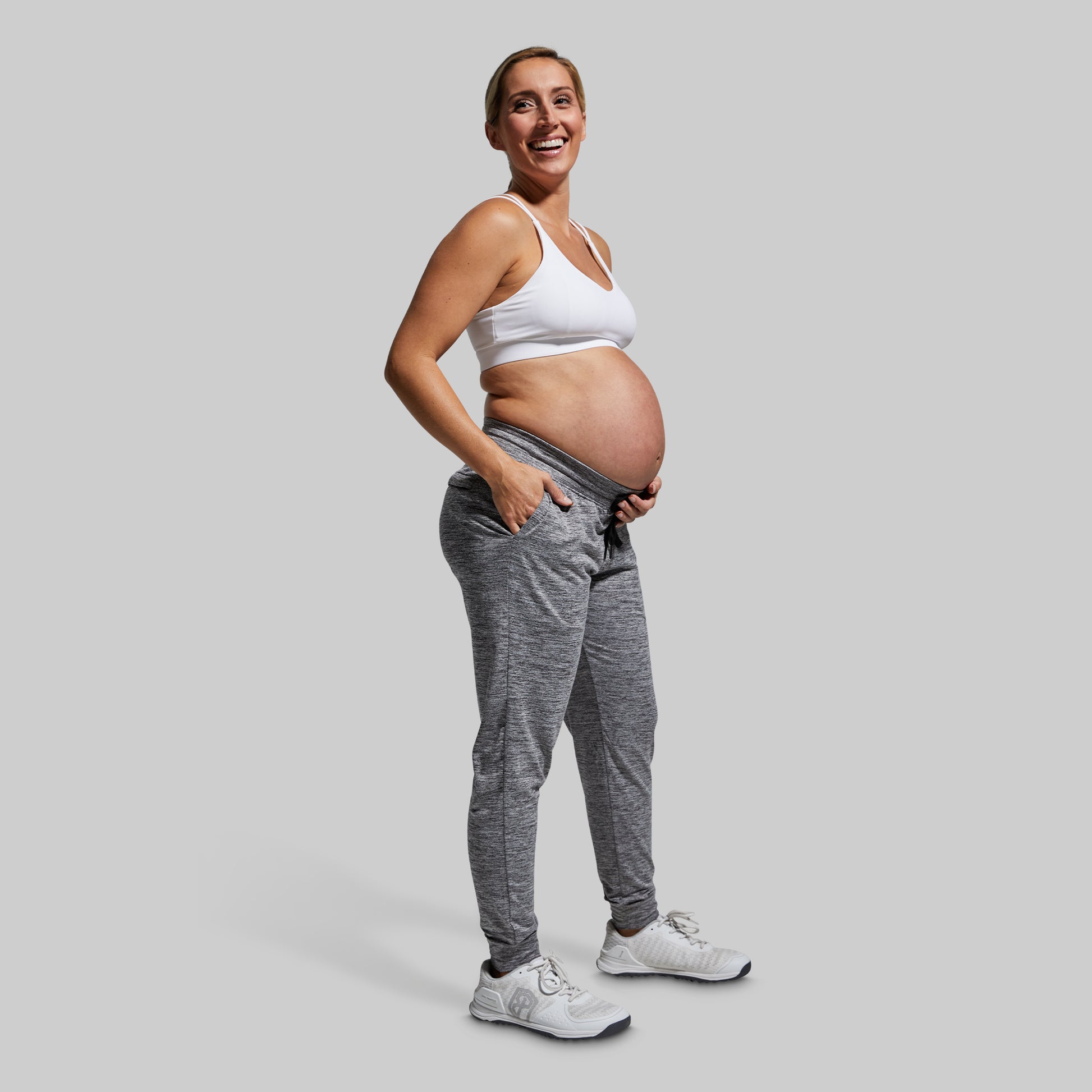 Pregnant woman wearing a white sports bra and gray jogger pants on a light gray background