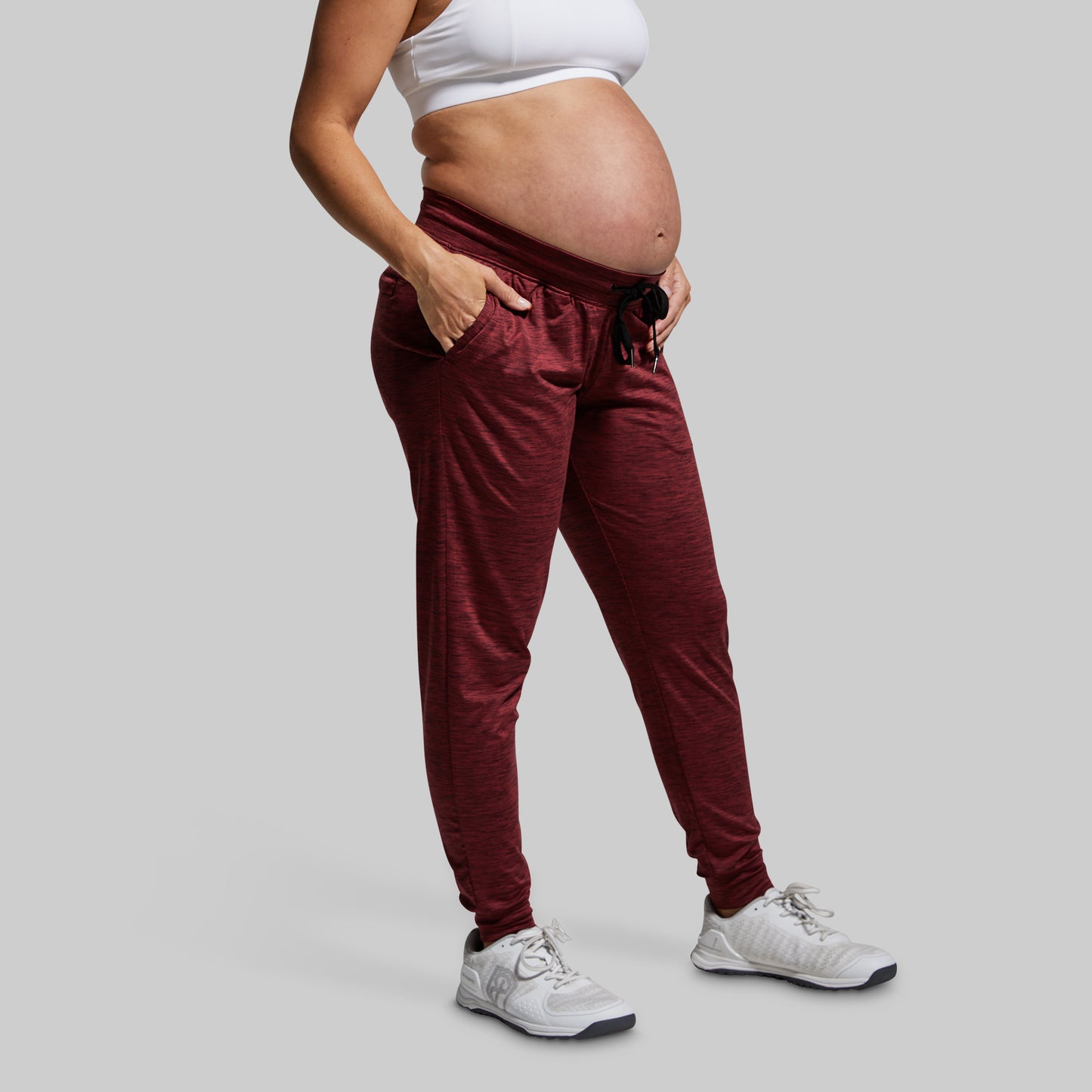 Pregnant person wearing maroon jogger pants and a white top on a gray background