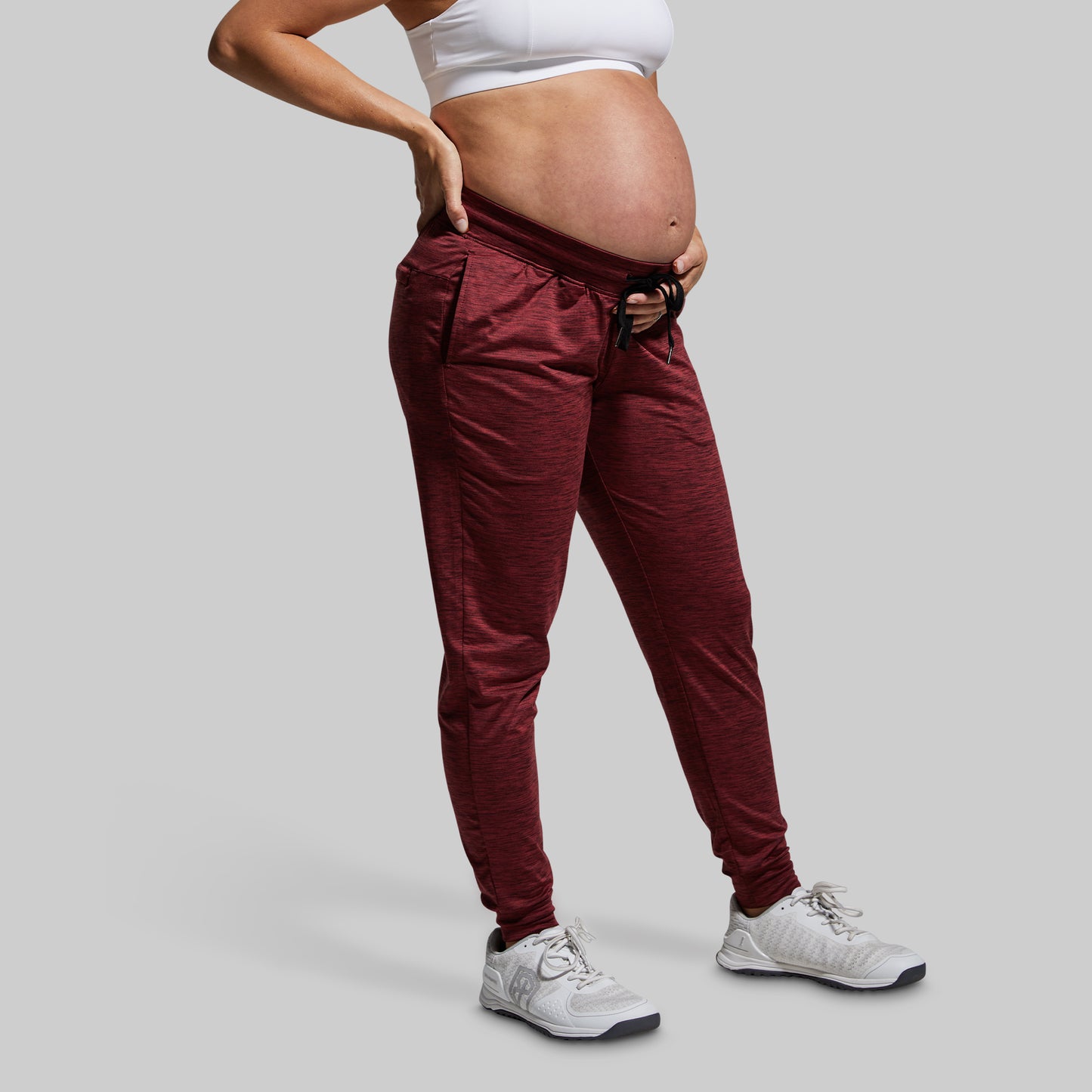 Pregnant woman wearing maroon joggers and white sneakers on a gray background