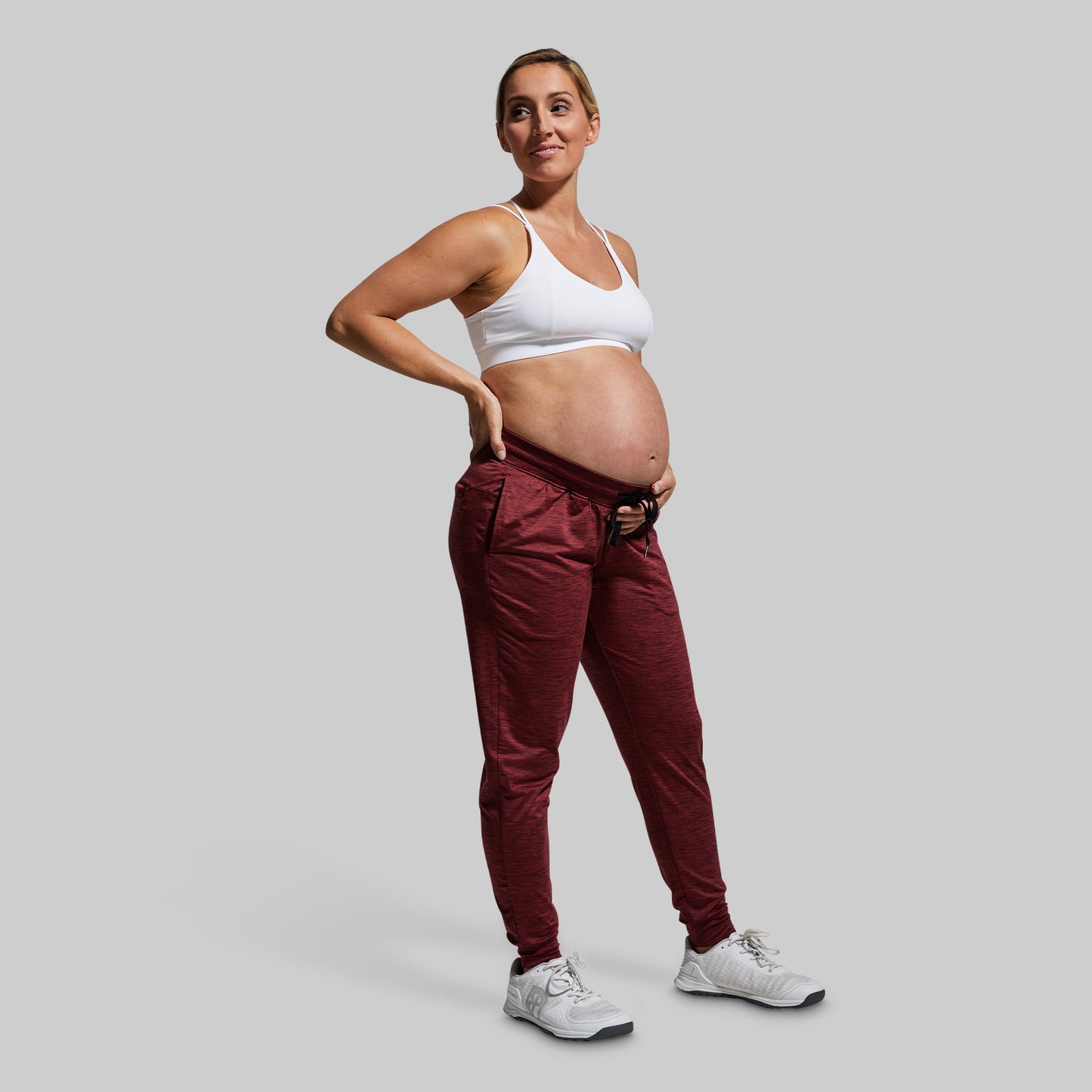 Pregnant woman wearing a white sports bra and maroon joggers on a gray background