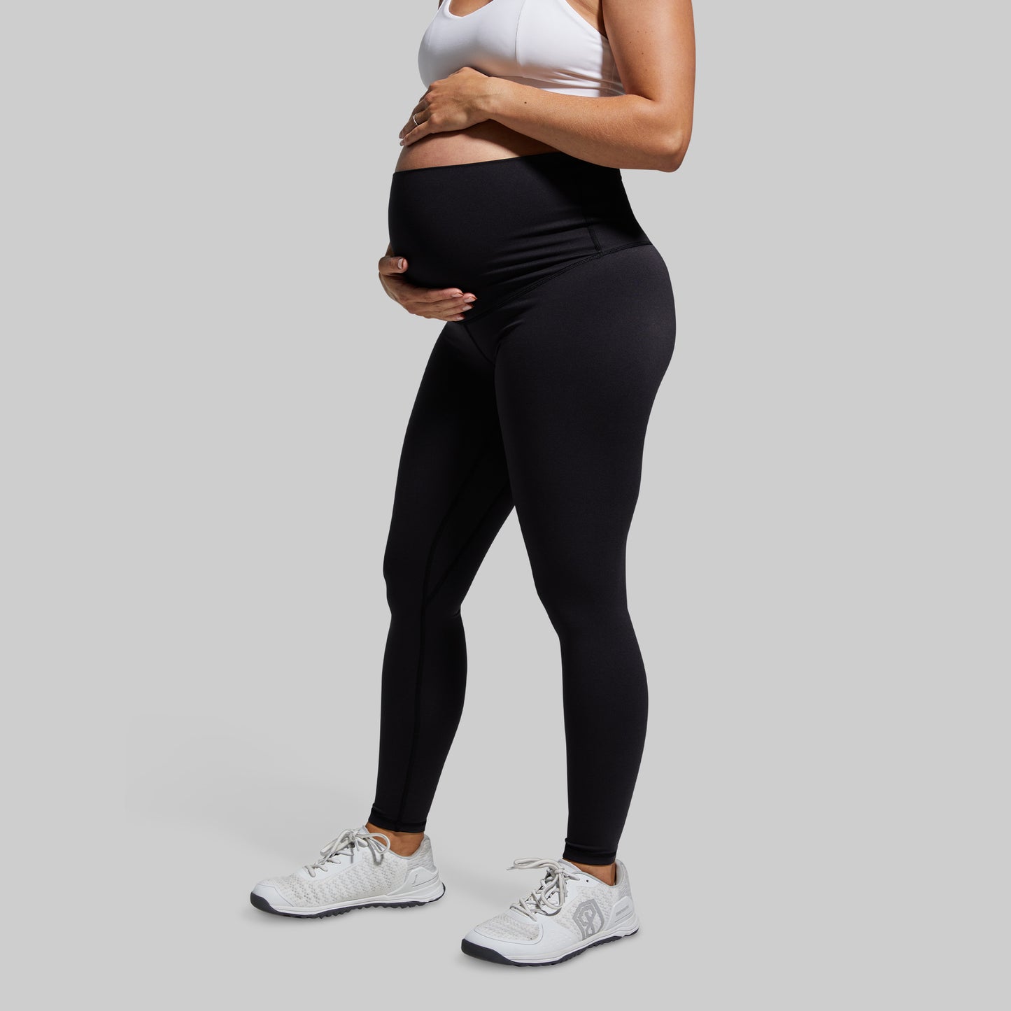 A pregnant woman wearing high-waisted black maternity leggings and white sneakers.