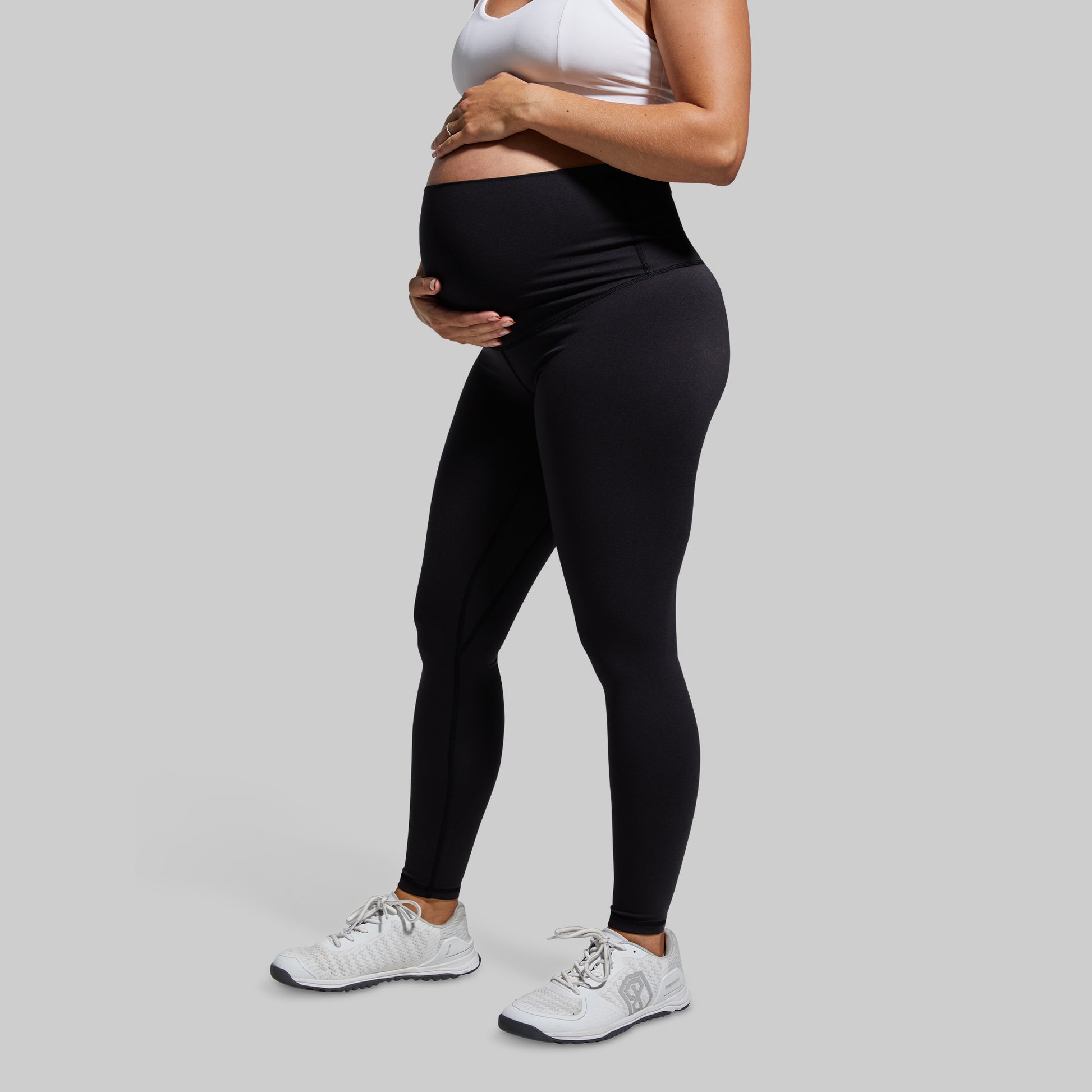 A pregnant woman wearing high-waisted black maternity leggings and white sneakers.