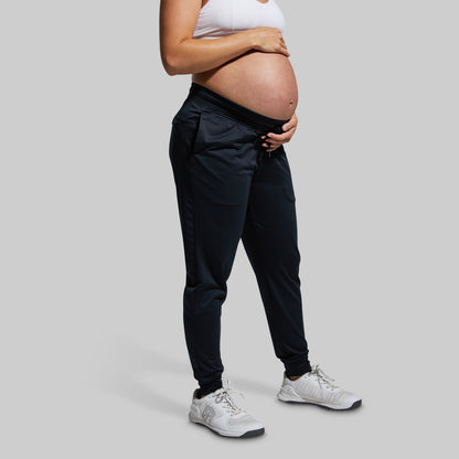 Pregnant person wearing black joggers and white sneakers on a gray background