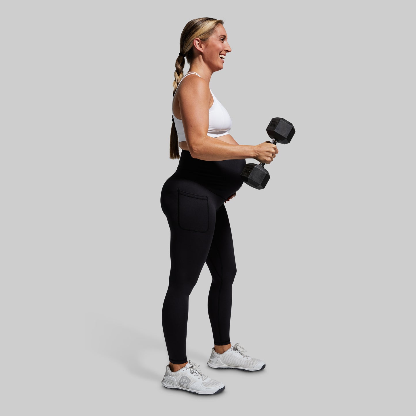 Pregnant woman exercising with a dumbbell wearing black leggings on a gray background
