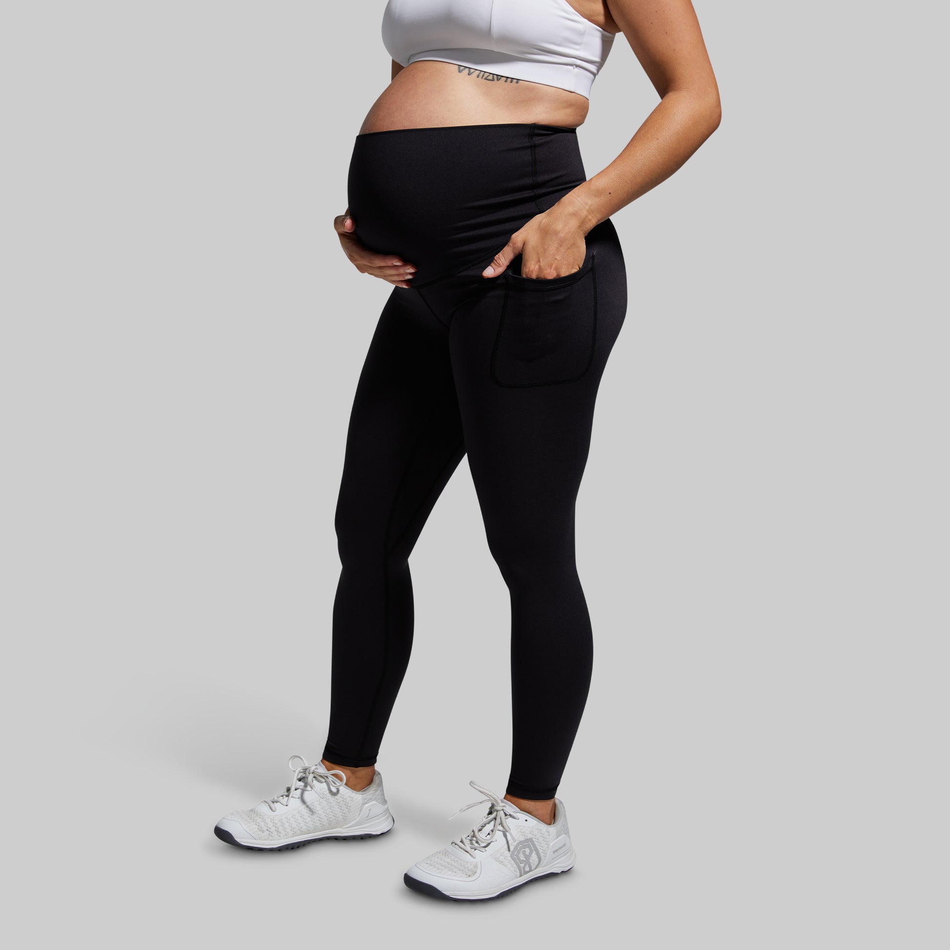 Person wearing black maternity leggings and a white sports bra on a gray background