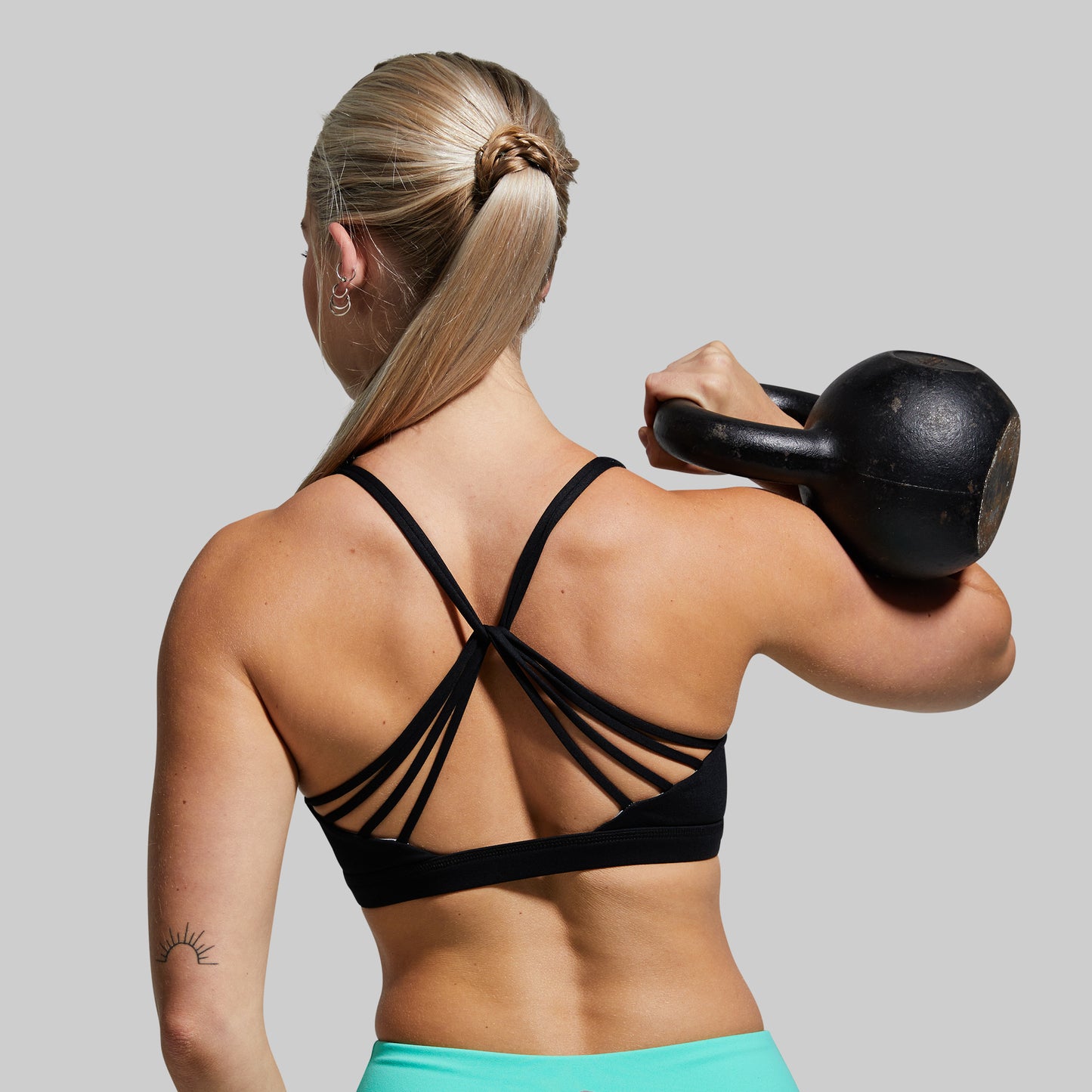 Woman wearing black sports bra and lifting a black kettlebell on a plain background