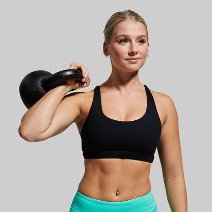 Woman wearing black sports bra and lifting a black kettlebell on a plain background