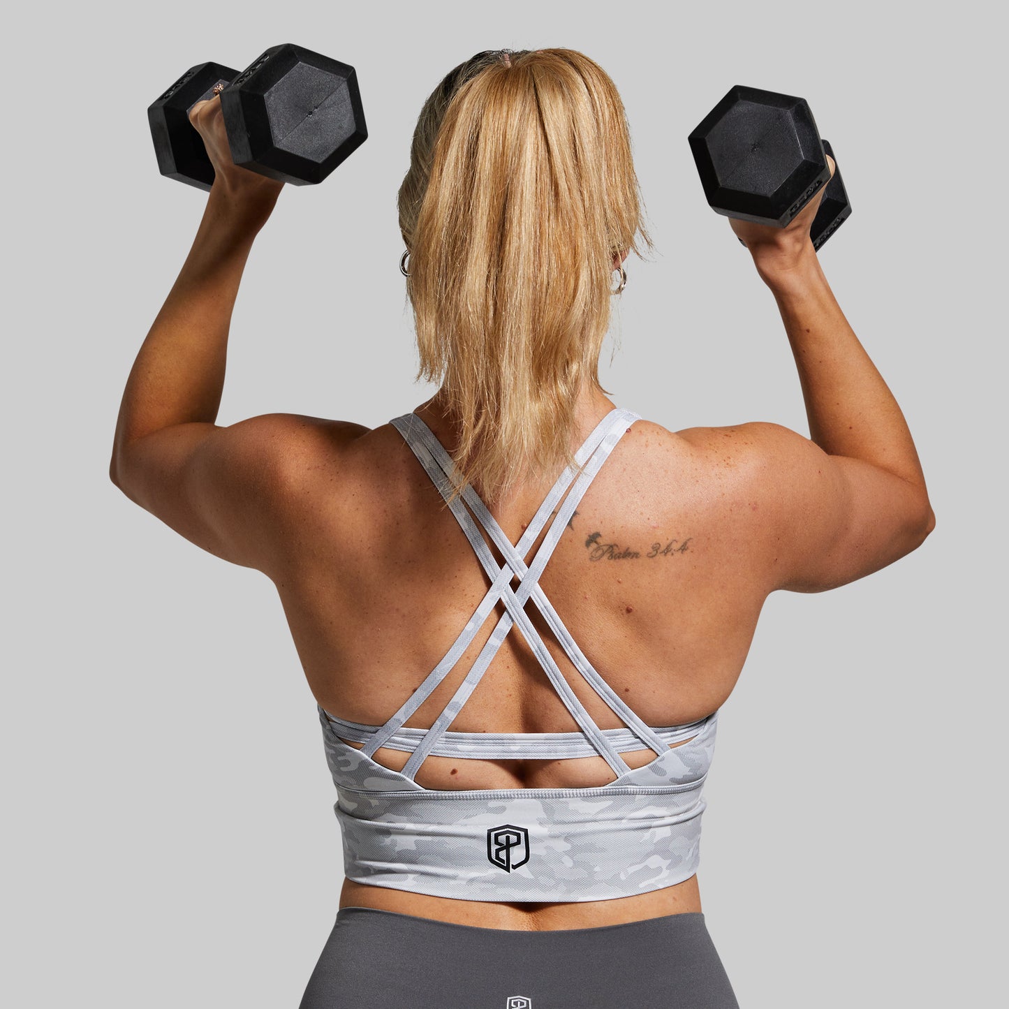 Woman lifting dumbbells from behind wearing a sports bra and leggings on a gray background