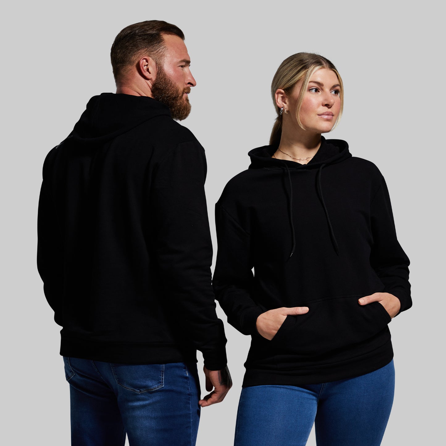 Man and woman wearing black hoodies and blue jeans against a gray background