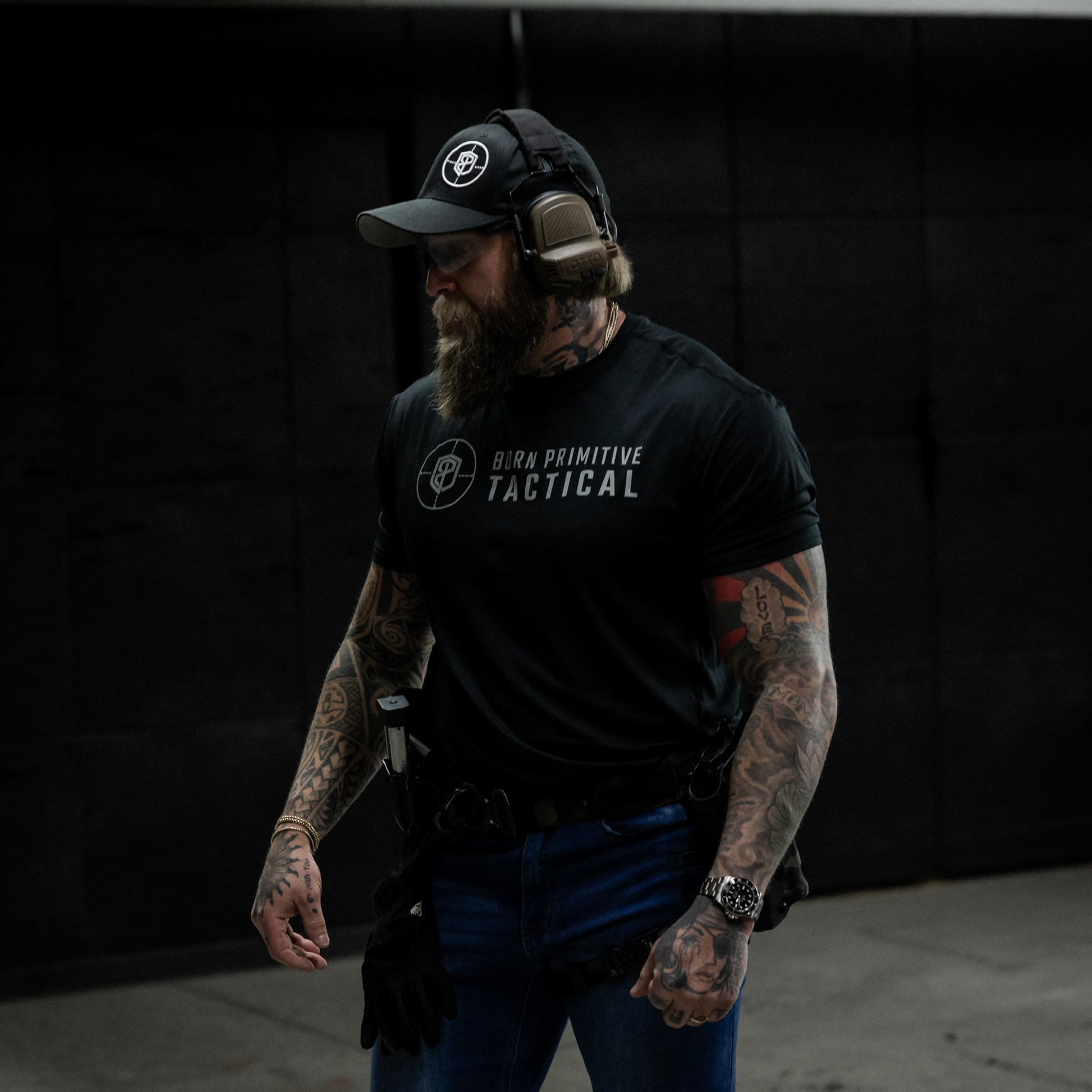 Man wearing a black 'Born Primitive Tactical' t-shirt with tactical gear against a dark background
