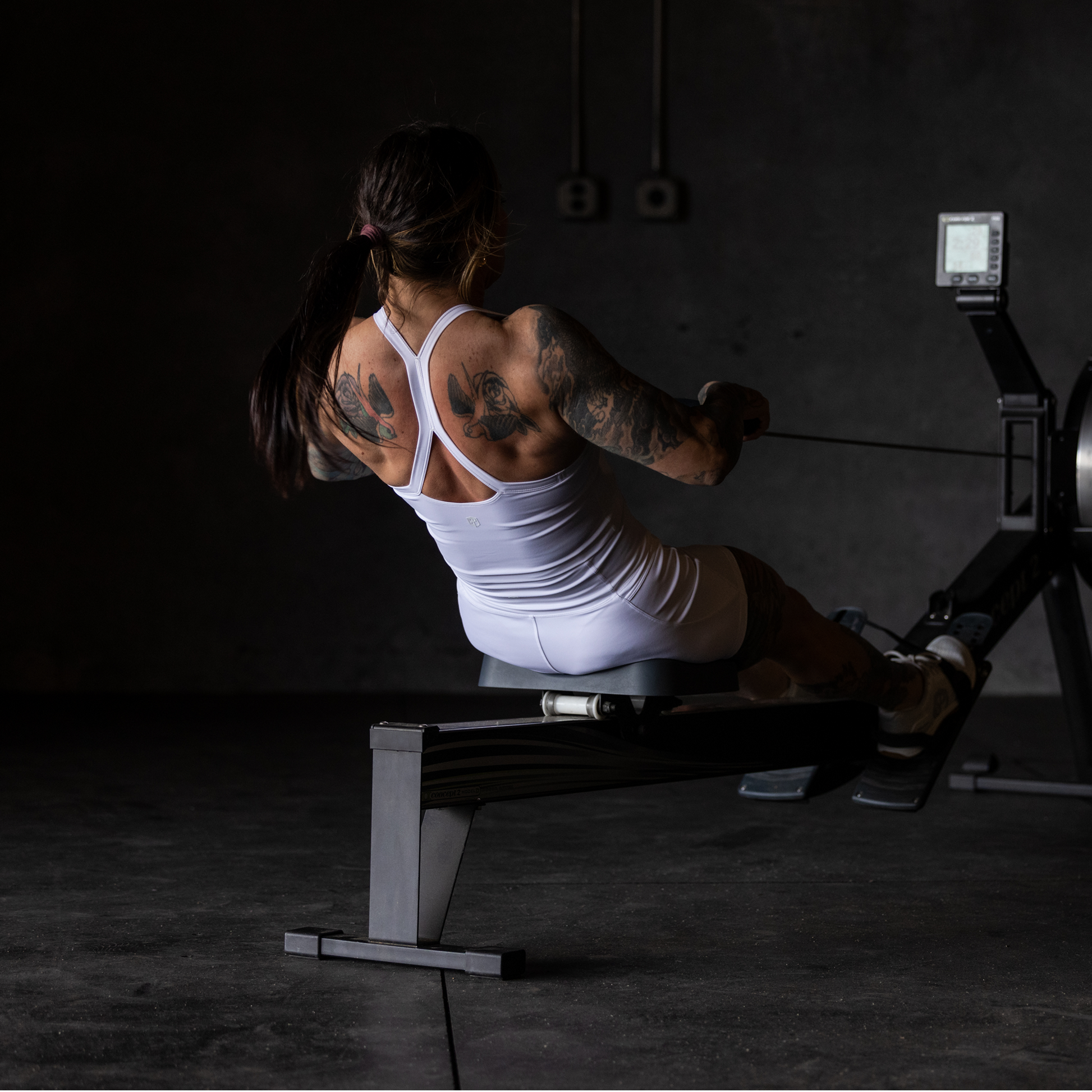 Person using a rowing machine in a dark indoor setting wearing a white sports bra.