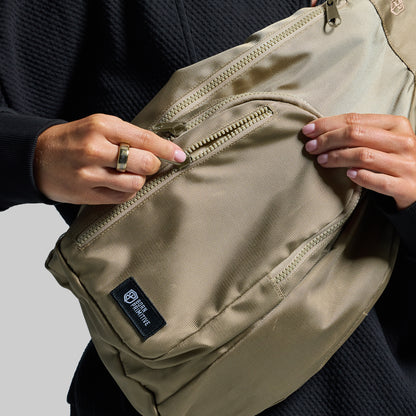 A close-up of a person wearing a beige crossbody bag with a black textured top. The person's hands are shown sliding the front zip pocket open, revealing a black brand label that reads 'BORN PRIMITIVE'.