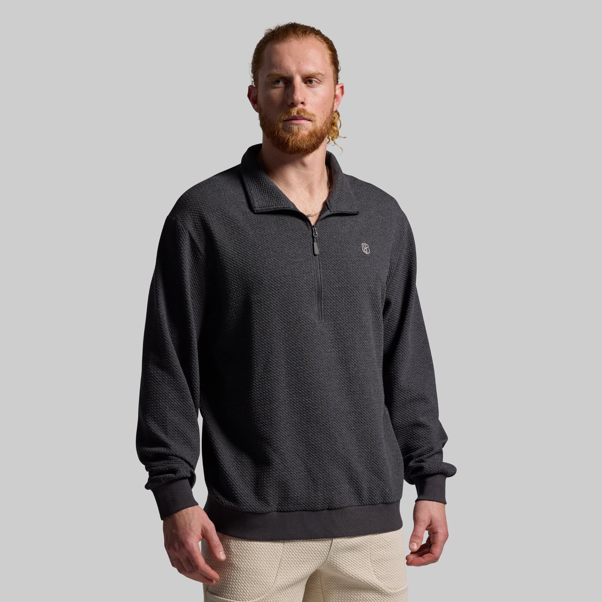 A man wearing a gray sweatshirt with a 1/4 zip front and a brand logo on the chest, paired with beige joggers.