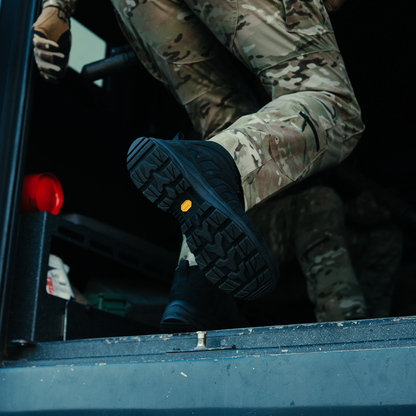 Person in camouflage uniform and boots standing next to a vehicle, with a focus on the boot.