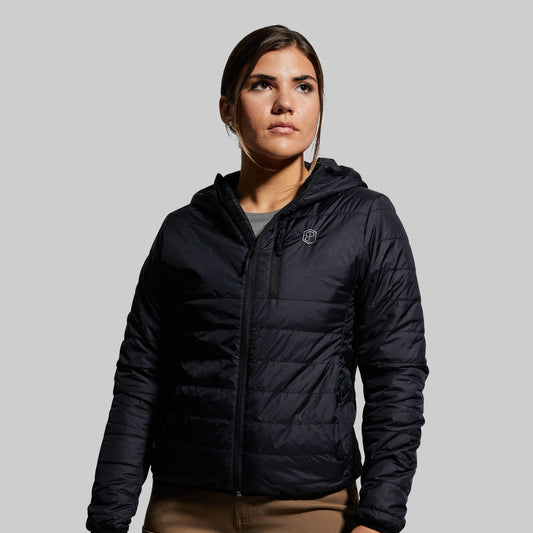 A woman wearing a black puffer jacket with a hood, standing against a plain background.