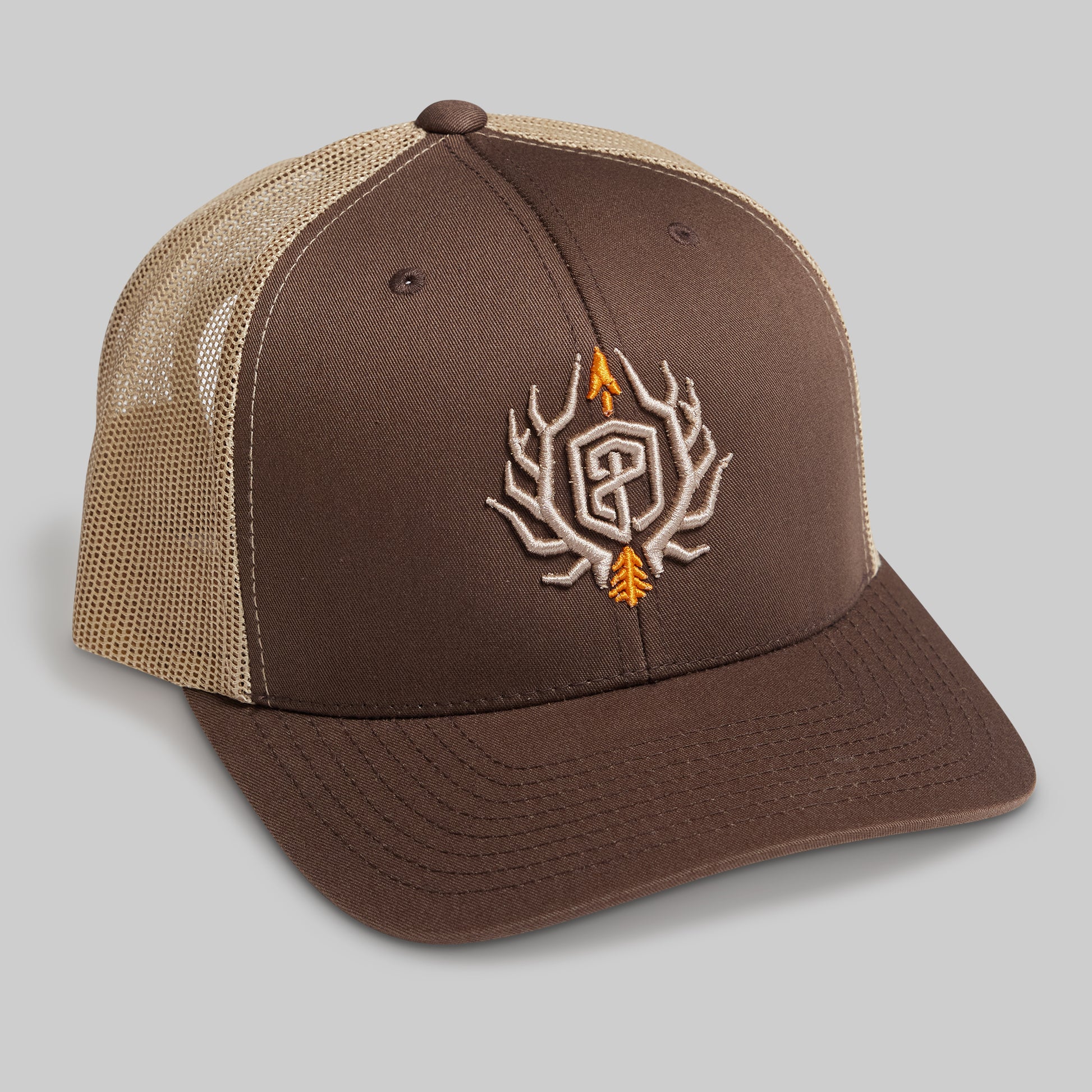 A brown and tan trucker-style cap with a brown front panel and brim, tan mesh back, and an embroidered antlered monogram logo on the front in brown, white, and orange.