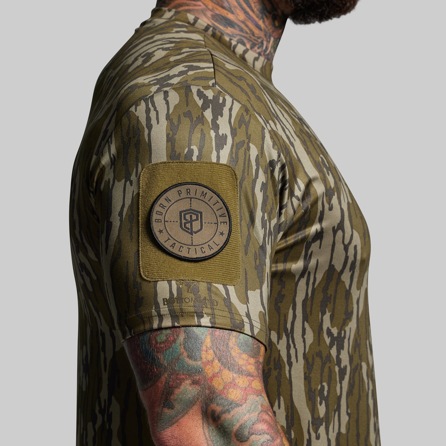 Person wearing a camouflage shirt with a visible brand patch on a gray background