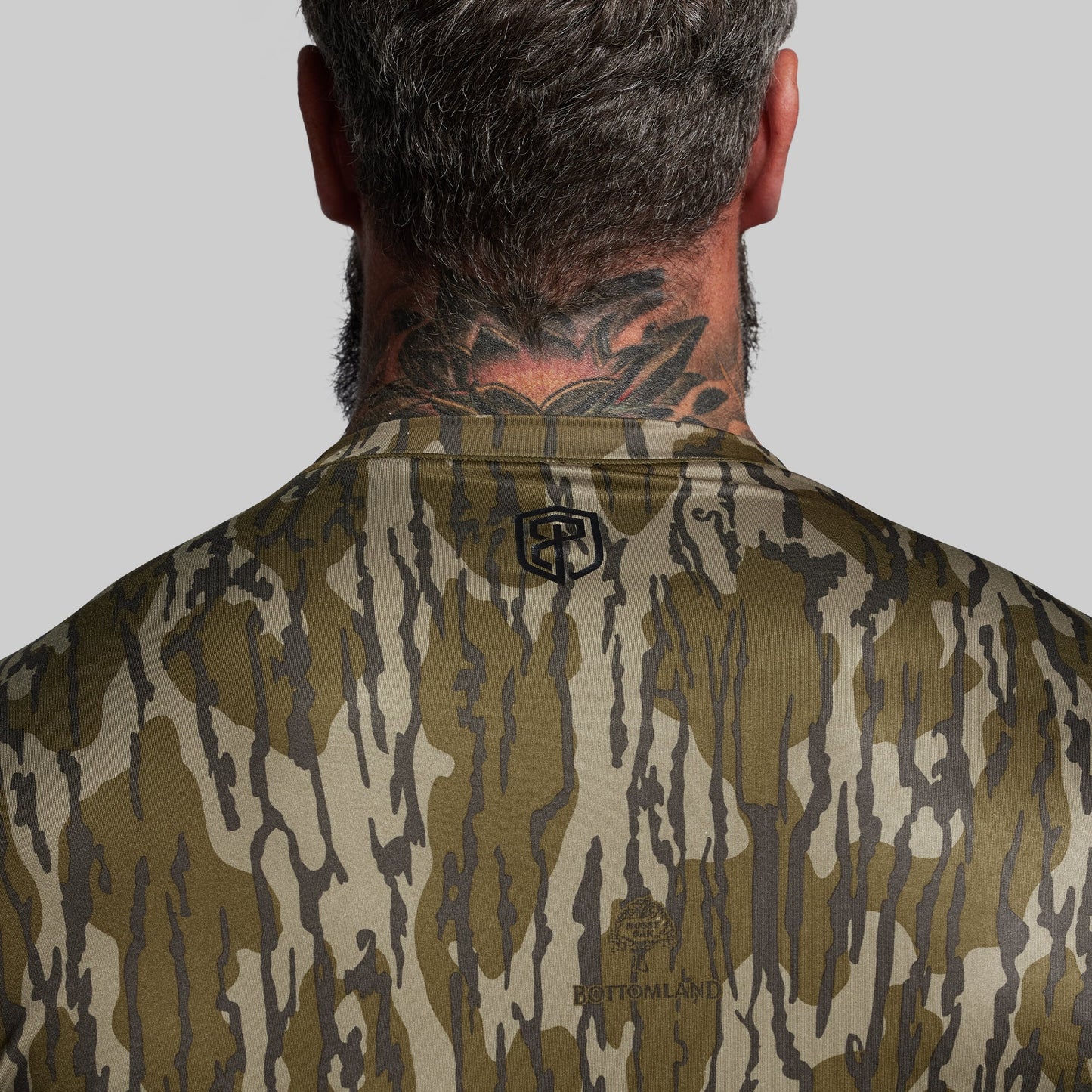 Person wearing a camouflage shirt with a brand logo on a plain background