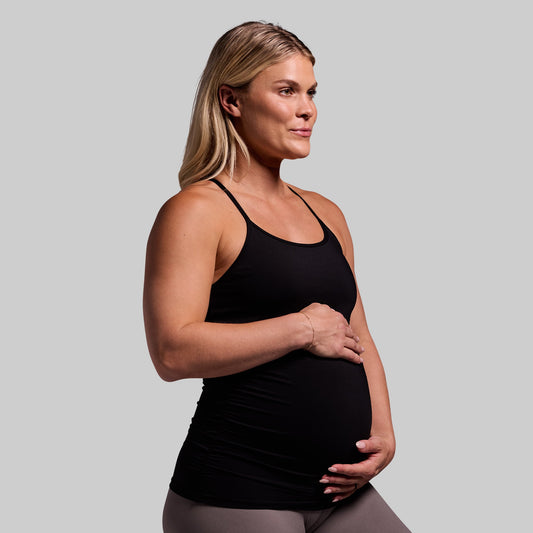 A pregnant woman wearing a black maternity drop shot tank top with a built-in bra layer, sleeveless design, and ruched sides.