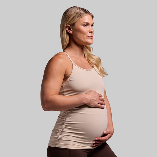 A pregnant woman wearing a beige maternity drop shot tank top with a built-in bra layer, sleeveless design, and ruched sides.