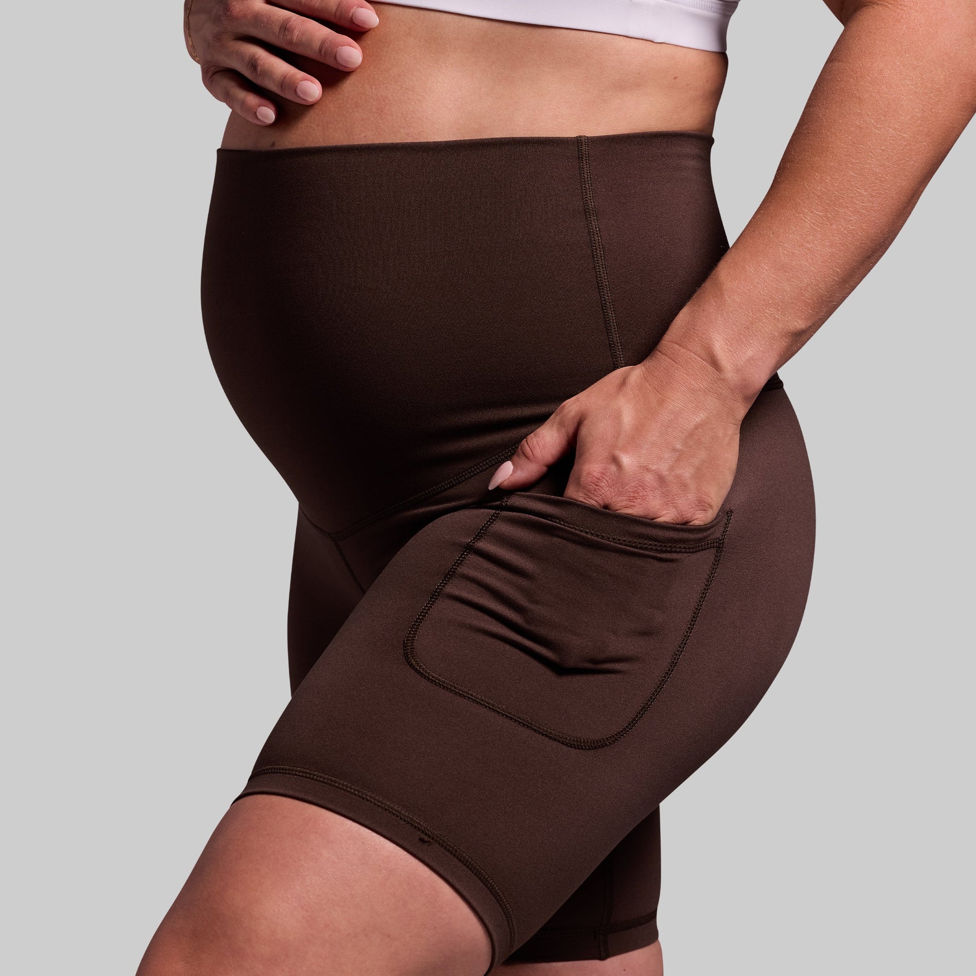 A pregnant woman wearing dark brown maternity biker shorts with a high waist and pockets.