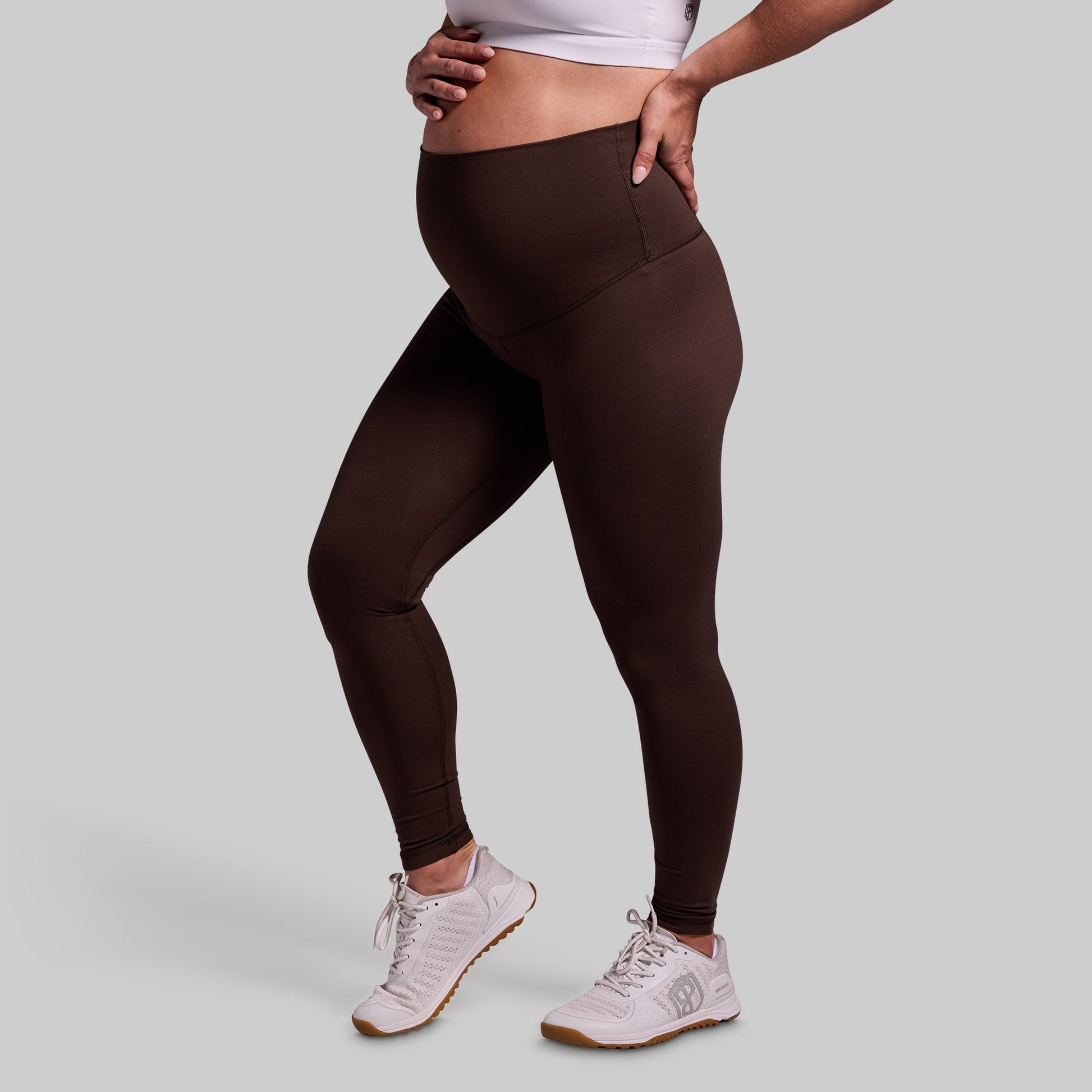 A pregnant woman wearing dark brown maternity leggings and white sneakers.