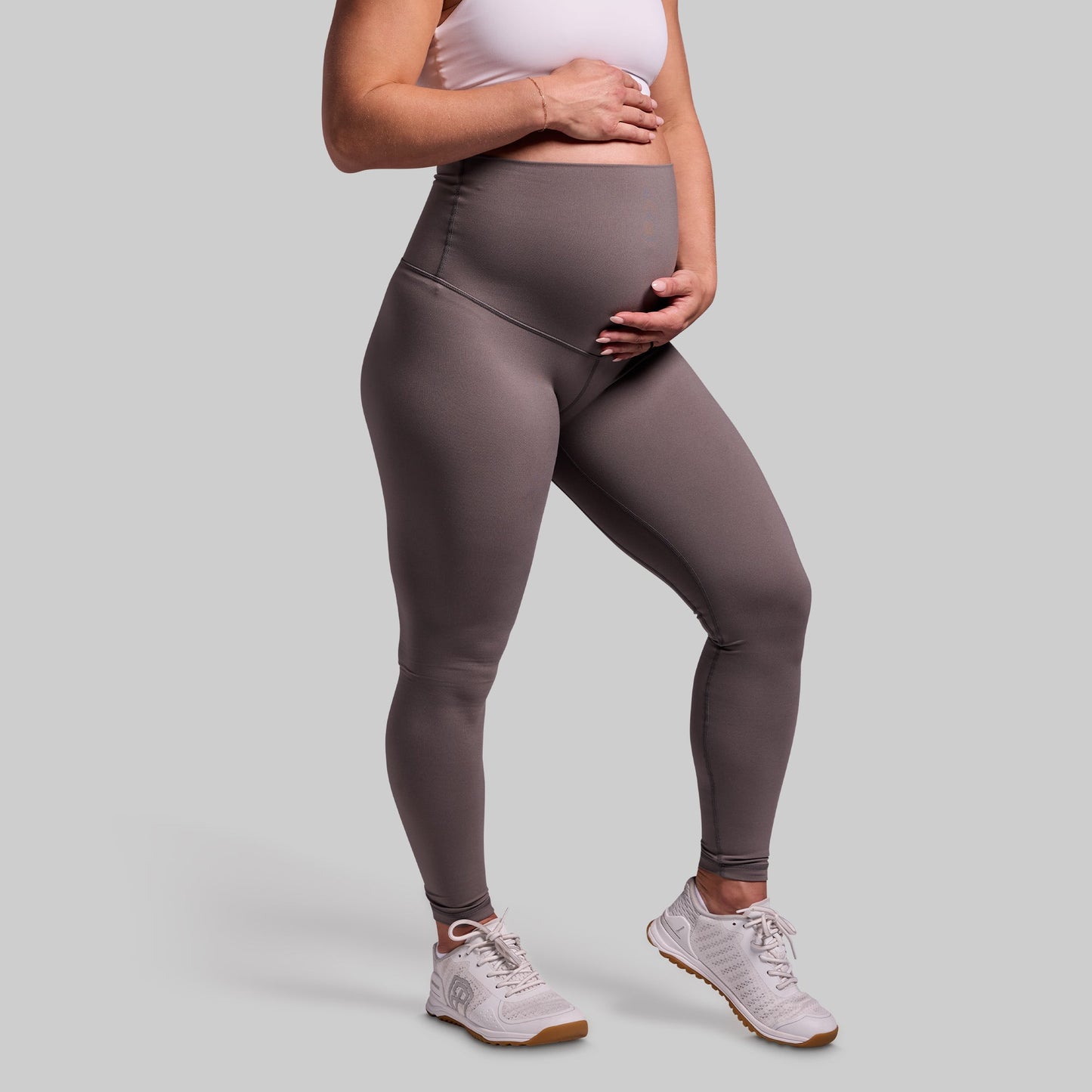 A pregnant woman wearing high-waisted grey maternity leggings and white sneakers.