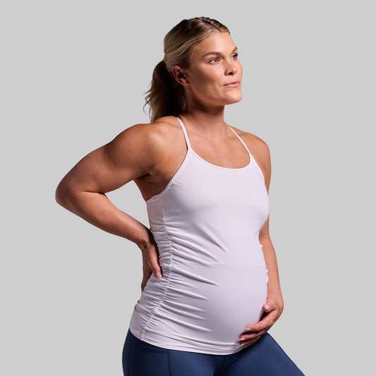A pregnant woman wearing a white maternity drop shot tank top with built-in bra layer, sleeveless design, and ruched sides.