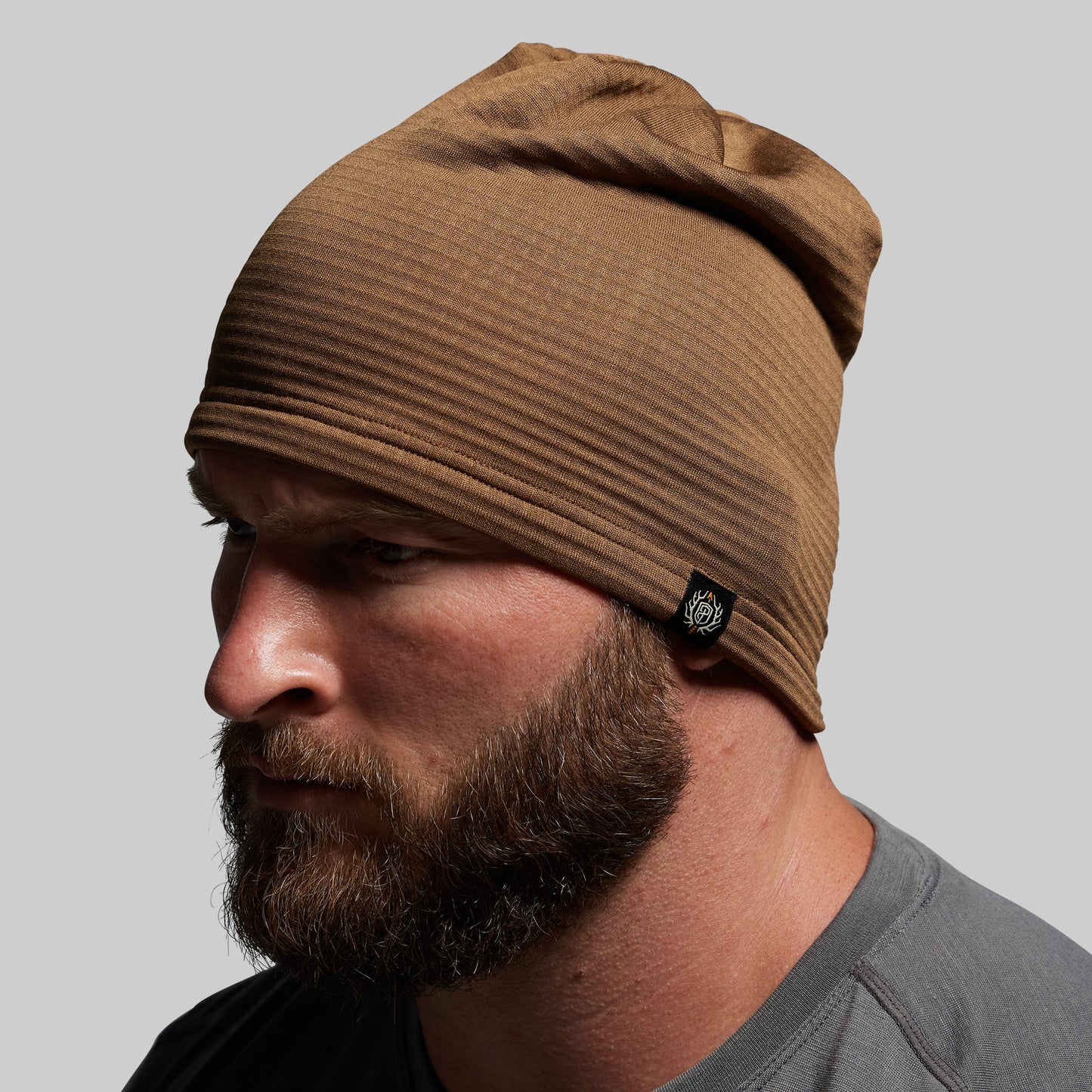 Man wearing a brown beanie with a brand logo on a gray background