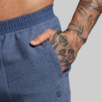 Person wearing blue shorts with a tattooed arm on a gray background