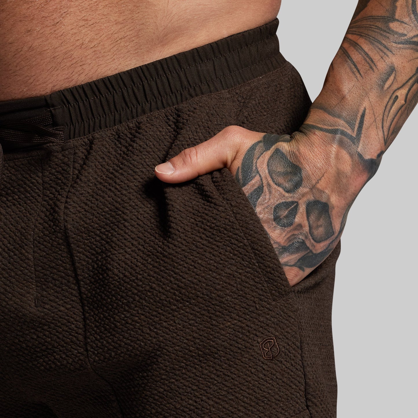 Brown textured shorts worn by a person with tattooed arm on a light gray background
