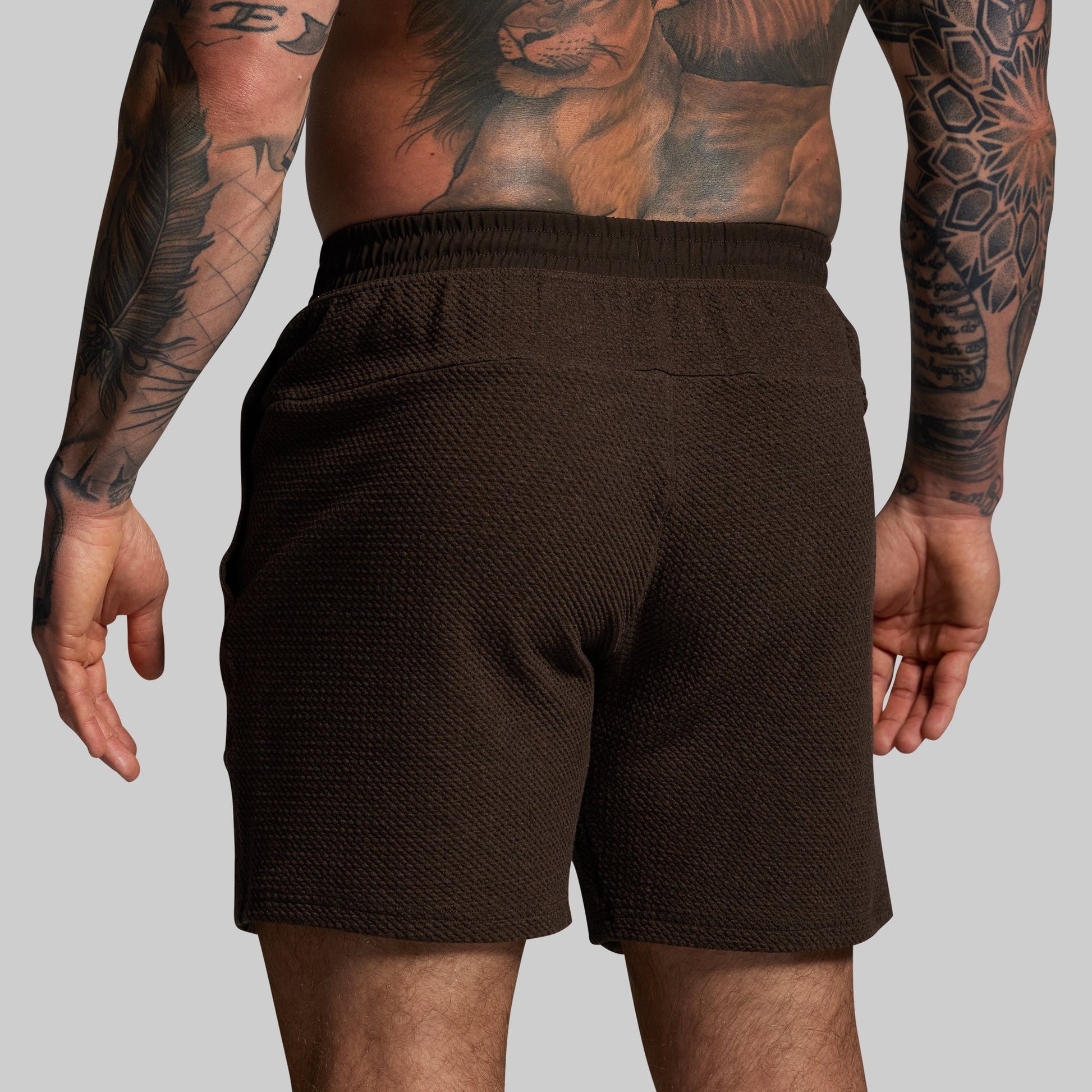 Person wearing brown shorts with tattoos on arms and legs against a gray background