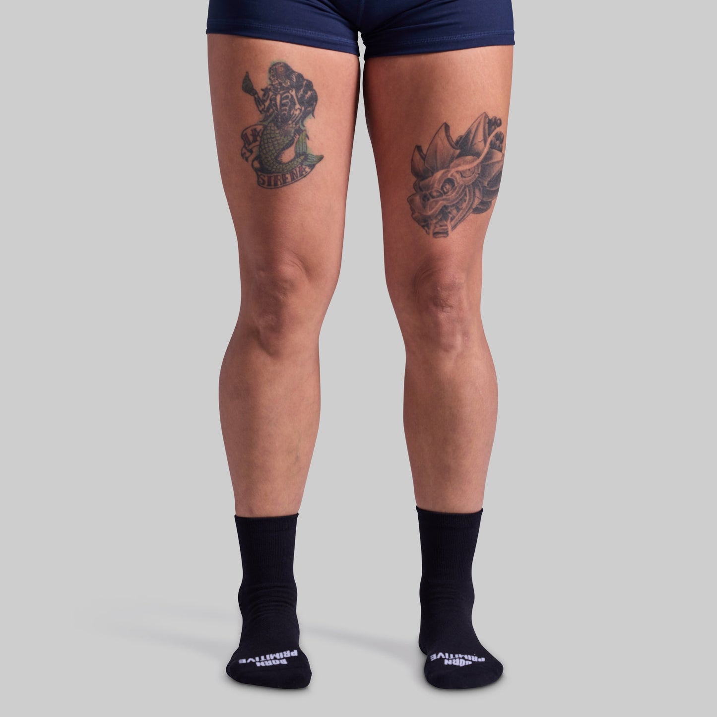 Person wearing navy blue shorts and black socks with visible tattoos on a gray background