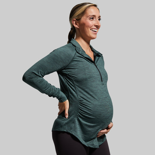 A pregnant woman wearing a long sleeve maternity top with a zip neck in a dark green color.
