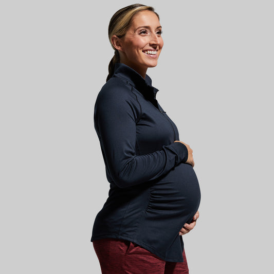 A pregnant woman wearing a long sleeve black maternity top with a zip neck.