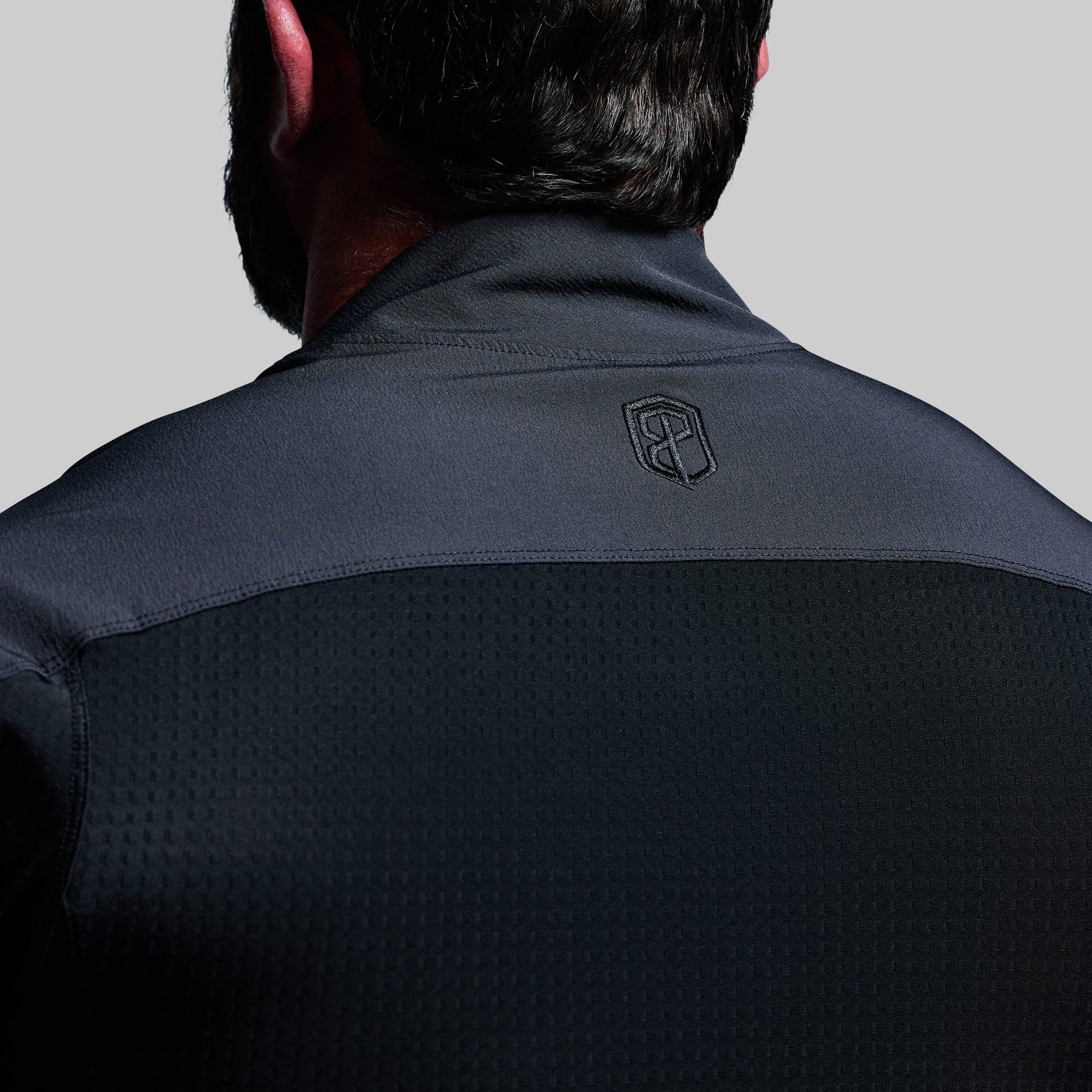 Back view of a person wearing a black tactical shirt with a logo on a gray background
