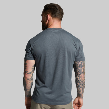 Man wearing a gray t-shirt with tattoos on arms against a plain background
