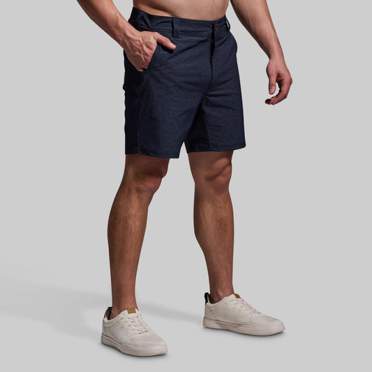 A man standing with one hand in a pocket, wearing navy-colored shorts and white sneakers.