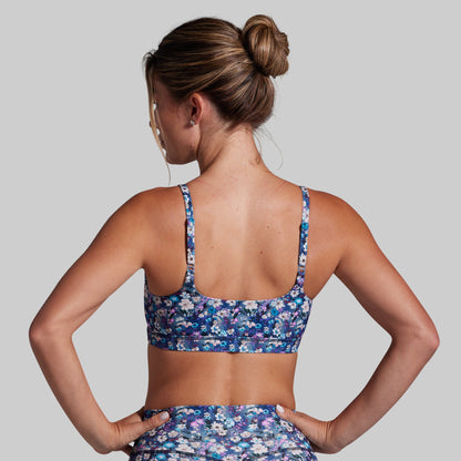 A woman shown from the back wearing a blue floral sports bra and matching high-waisted bottoms against a solid gray studio background.