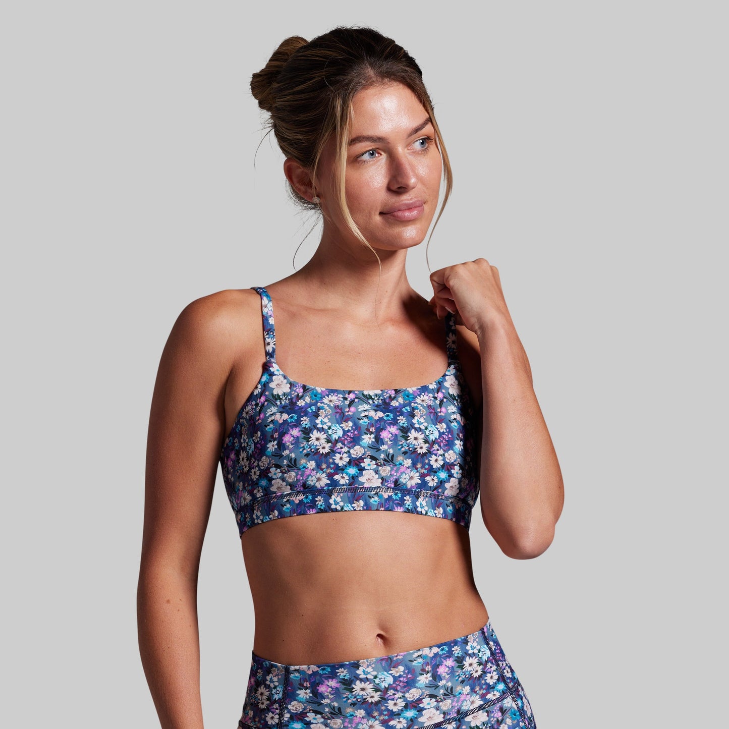 A woman modeling a matching floral print activewear set, featuring a blue sports bra and high-waisted bottoms with a purple, blue, and white floral pattern, set against a solid gray background.