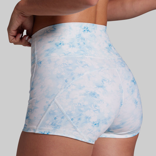 Close-up back view of a model wearing high-waisted athletic shorts with a light blue and white tie-dye pattern, set against a solid gray background.