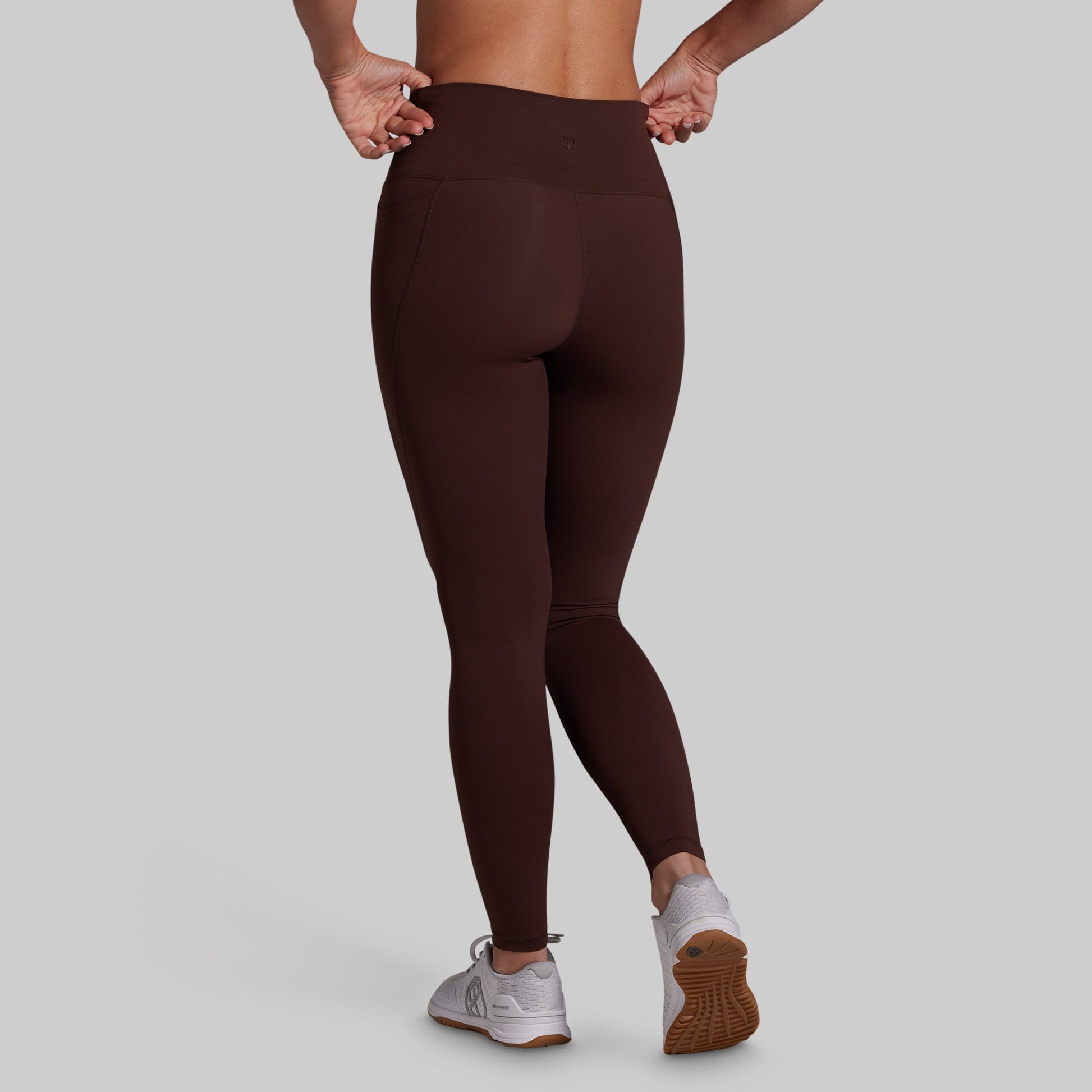A person shown from the back wearing dark brown high-waisted leggings and white athletic sneakers against a light gray studio background.