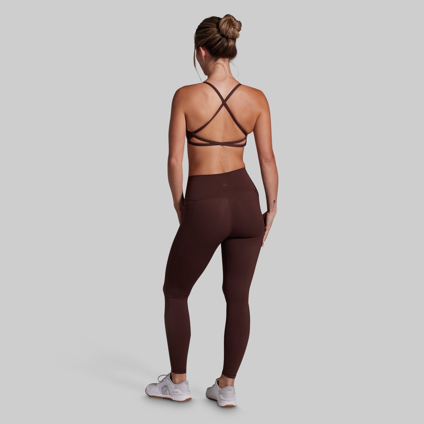 A woman shown from the back wearing dark brown athletic leggings and a matching sports bra, standing against a solid light gray background.
