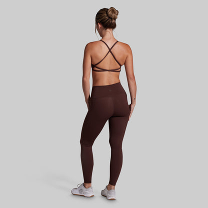 A woman shown from the back wearing dark brown athletic leggings and a matching sports bra, standing against a solid light gray background.